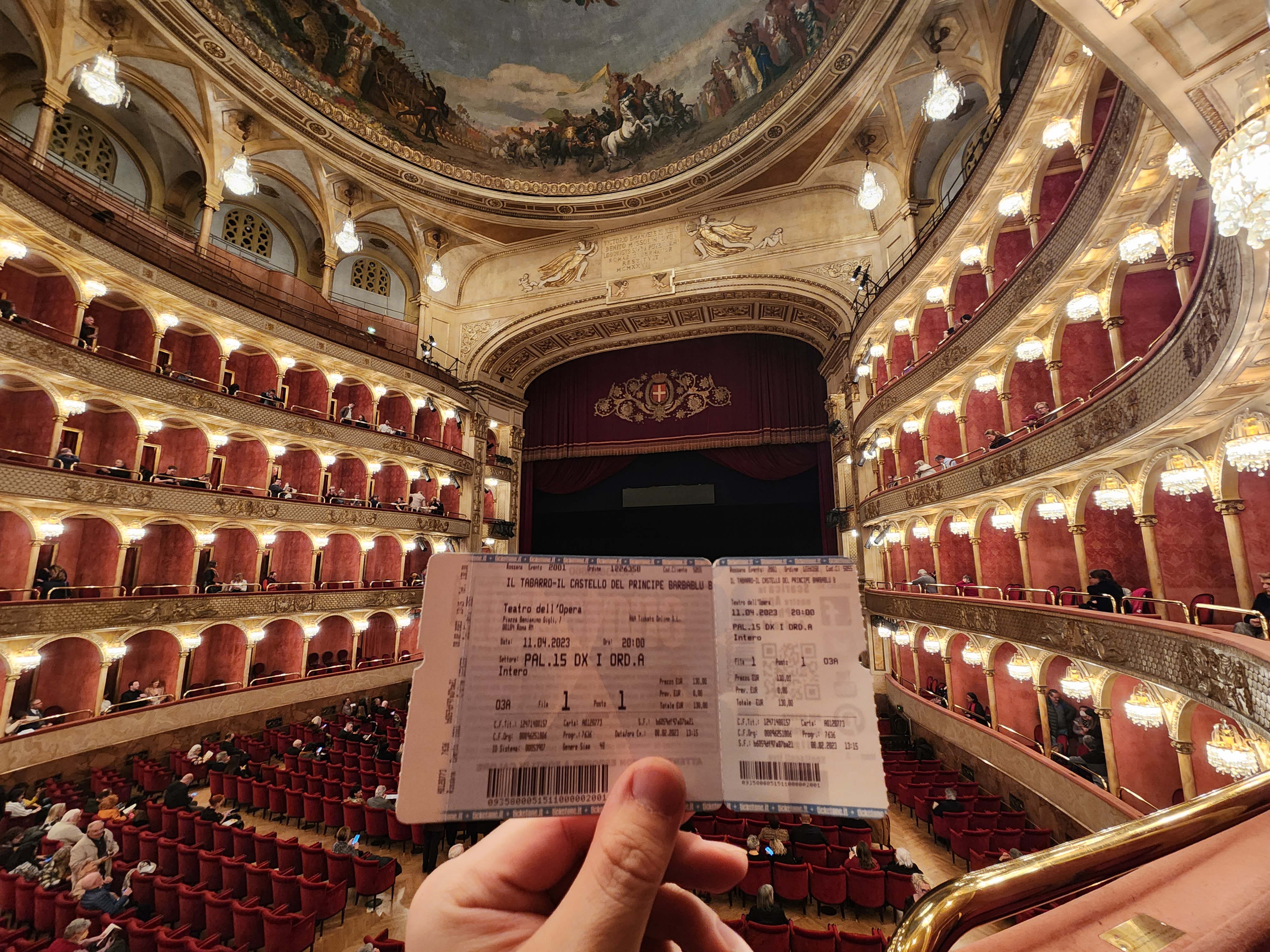 A hand holding an opera ticket inside an ornate theatre.